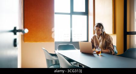 Eine junge Frau mit Kopfhörern arbeitet an ihrem Laptop in einem hellen, modernen Bürobereich. Fokussiert und produktiv verkörpert sie ein professionelles Umfeld Stockfoto