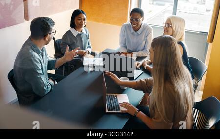 Eine Gruppe von Geschäftsleuten unterschiedlichen Alters nimmt während einer Besprechung in einem Büro an einem Gespräch Teil. Mehrere Laptops und Notizen werden auf den Tisch gestellt, re Stockfoto