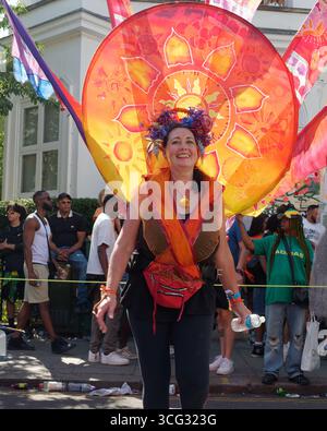Frau im Kostüm mit großer orangener Scheibe lächelt während des Notting Hill Karnevals mit Zuschauern dahinter, London. England, 25. August 2025 Stockfoto