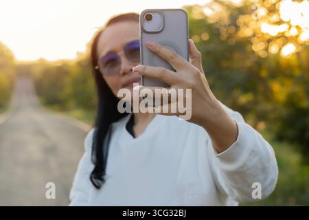 Frau, die Selfie mit Smartphone macht, auf der Straße im wunderschönen Wald bei Sonnenaufgang. Selektiver Fokus. Stockfoto