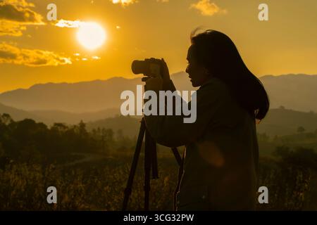 Silhouette einer Fotografin, die ein Foto bei Sonnenuntergang auf einem Berg macht. Seitenansicht. Selektiver Fokus. Stockfoto