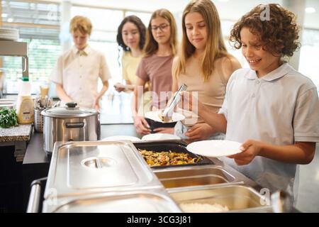 Verschiedene Gruppen von Schülern stehen in einer gut beleuchteten Schulcafeteria zum Mittagessen an, genießen frisch zubereitete Speisen und freundliche Interaktionen, wobei ein po betont wird Stockfoto