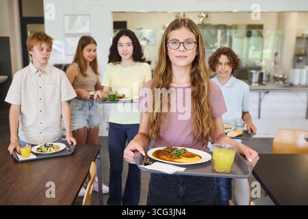Die Schüler treffen sich in einer Cafeteria, halten Tabletts mit Mahlzeiten und Getränken bereit und genießen die gemeinsame Mittagszeit. Stockfoto
