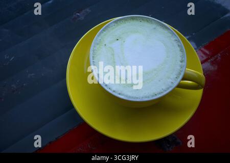 Matcha-Grüntee-Latte mit Latte Art, serviert in einer gelben handgefertigten Keramikschale auf einem Vintage-Metalltisch. Stockfoto