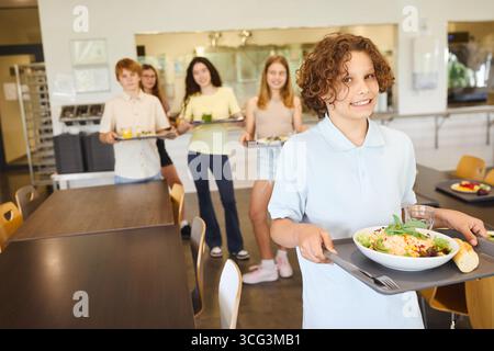 Gruppe von Schülern in einer Schulcafeteria mit Tabletts mit gesunden Nahrungsoptionen. Die Szene bietet eine freundliche Atmosphäre, abwechslungsreiche Mahlzeiten und eine saubere Atmosphäre Stockfoto