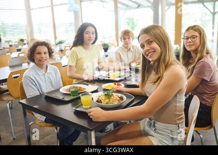 Eine Gruppe von fünf Teenagern lächelt beim gemeinsamen Mittagessen in einer gut beleuchteten Schulcafeteria. Sie sitzen an einem Tisch und genießen eine Auswahl an Mahlzeiten Stockfoto