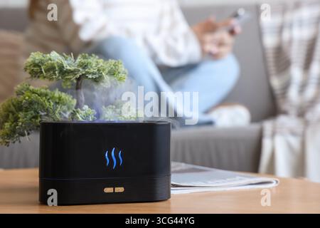Luftbefeuchter mit Bonsaibaum auf Tisch gegen Frau mit Handy im Wohnzimmer, Nahaufnahme Stockfoto