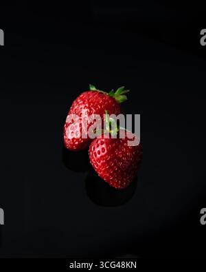 Nahaufnahme zweier frischer Erdbeeren mit grünen Stielen auf reflektierender schwarzer Oberfläche. Die roten Erdbeeren heben sich vor dem dunklen Hintergrund hervor, hochglänzend Stockfoto
