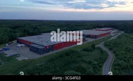 Blick in die Abenddämmerung auf ein modernes Logistiklager mit Laderampen und Sattelzügen, mit nachhaltiger Architektur, die in einen grünen Wald integriert ist Stockfoto