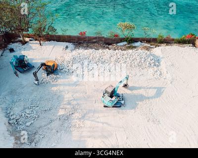 Baustelle mit Baggern, die weiße Felsen in der Nähe des türkisfarbenen tropischen Meeres bewegen, aus der Vogelperspektive Stockfoto
