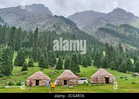 Kirgisistan, Cholpon-ATA - 16. Juli 2025: Traditionelle Jurten in einem grünen Tal der Barskoon-Schlucht, umgeben von Kiefernwäldern und felsigen Berggipfeln Stockfoto