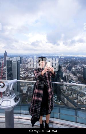 Wunderschöne elegante Frau, die im Winter herbstlich modisches Outfit trägt, während sie Selfie auf der Spitze des Hauptturms macht und den Blick auf den Wolkenkratzer genießt Stockfoto