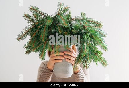 Eine junge Frau in einem warmen, gemütlichen Pullover mit einer rustikalen Tonvase gefüllt mit frisch geschnittenen Nadelzweigen aus grüner Tanne, isoliert auf weiß Stockfoto