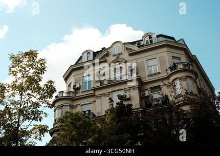 Elegantes historisches Gebäude mit Balkonen, Fenstern und Klimaanlagen, eingefangen vor blauem Himmel mit Baumblättern im Vordergrund. Belgrad, Serbien. Stockfoto