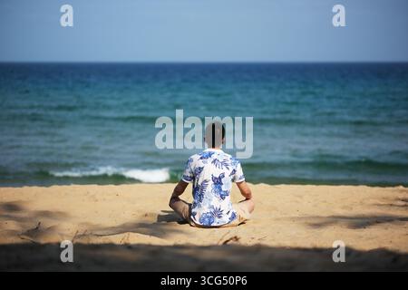 Mann, der am Strand sitzt und auf den Ozean blickt. In Sanya, Hainan, China Stockfoto