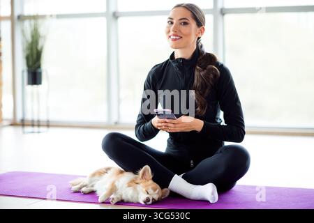 Die junge Frau mit ihrem Hund genießt ein ruhiges Indoor-Workout auf einer Yogamatte, während sie ein Telefon hält Stockfoto