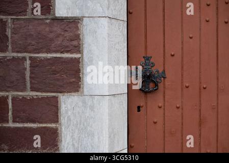 Eine detaillierte Ansicht einer strukturierten, braunen Holztür mit einem verzierten schwarzen Metallklopfer an einer Ziegelsteinmauer. Altes dekoratives Schloss an einem alten Stockfoto
