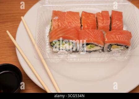 Sushi-Brötchen mit frischem Lachs, Avocado und Frischkäse, serviert in einem durchsichtigen Plastikbehälter auf Teller, begleitet von Essstäbchen und Sojasauce. Eine App Stockfoto