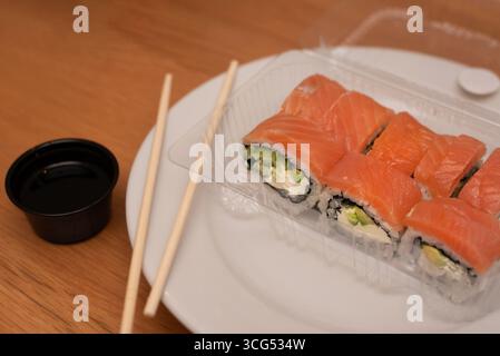 Frische Sushi-Brötchen mit Lachs, Avocado und Frischkäse werden zusammen mit Sojasauce und Essstäbchen in einem praktischen Behälter zum Mitnehmen serviert Stockfoto
