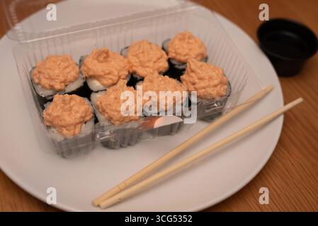 Köstliche Sushi-Brötchen mit einer cremigen, scharfen Sauce in einer durchsichtigen Verpackung, serviert mit Essstäbchen auf einem weißen Teller auf einem Holzteller Stockfoto