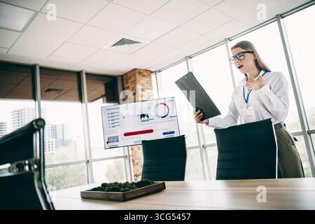 Junge Frau, die sich bei einer Firmensitzung mit einem Clipboard mit Hingabe in einem modernen Büro präsentiert Stockfoto