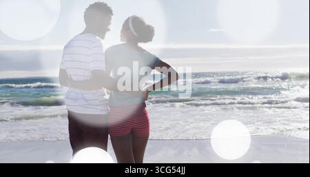 Stehendes afroamerikanisches Paar mit Hüften, mit Blick auf das ruhige Meer am Ufer unter hellem Himmel Stockfoto