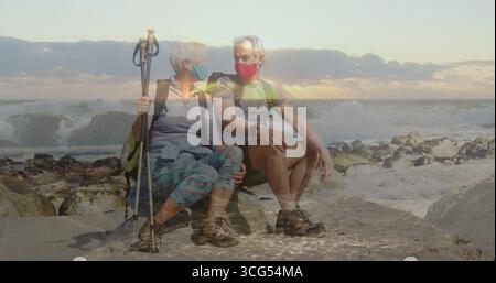 Sitzendes Seniorenpaar mit Wanderausrüstung und Masken an der felsigen Küste, mit Rucksäcken und Stangen Stockfoto
