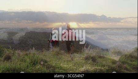 Spazieren Sie zu zweit Hand in Hand durch Küstengräser auf dem Weg bei Sonnenuntergang, Trekkingstöcke, Tagesrucksäcke Stockfoto