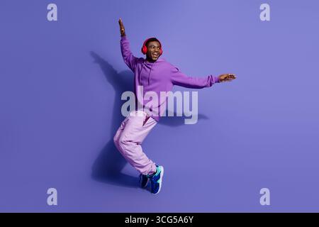 Energiegeladener junger Mann in leuchtendem lila Outfit, lächelt und tanzt mit dynamischer Pose vor farbigem Studiohintergrund Stockfoto