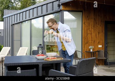 Ein Mann mittleren Alters steht an einem Terrassentisch des kleinen Hauses und beißt spielerisch mariniertes Schweinefleisch auf Spieß Stockfoto