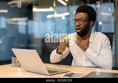 Mann in einer hellen Büroumgebung mit einem Rachenspray zur Linderung von Schmerzen. Das Bild vermittelt Gesundheitsbedenken, Medikamentenkonsum und Szenarien am Arbeitsplatz. Stockfoto