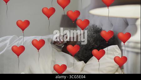 Eine Frau in weißem Pyjama, die im Schlafzimmer auf dem Bett liegt, mit roten Herzballons Stockfoto