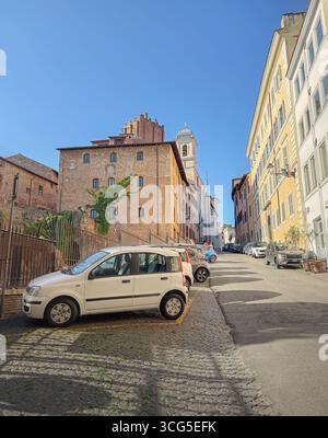 Kopfsteinpflasterstraße in Rom mit einer Mischung aus geparkten Autos und historischen Gebäuden. Die Straße führt bergauf und zeigt einen Glockenturm im Hintergrund und einen Stockfoto