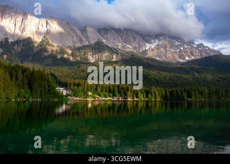 Wunderschöner Alpensee mit grünem Wald und Hütten unter hoch aufragenden felsigen Bergen mit dramatischen Wolken. Ein friedliches und landschaftlich reizvolles Reiseziel Stockfoto