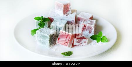 Türkisches Vergnügen. Lokum, bunte, gelatineartige Süßigkeiten mit Geschmack, bedeckt mit Puderzucker, serviert auf einem Teller auf einem weißen Tisch. Minzblätter Dekoration. Stockfoto