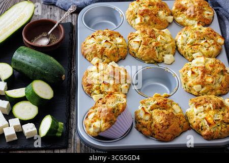 Nahaufnahme von herzhaften Zucchini Feta Muffins in Muffinform auf rustikalem Holztisch mit Küchentuch und Rohstoffen, horizontaler Blick von oben, nicht AI Stockfoto