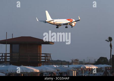 Larnaca, Zypern - 26. Juli 2025: Weißes und oranges Passagierflugzeug landet über dem Strand von Mackenzie mit Sonnenschirmen und Rettungsschirmen Stockfoto