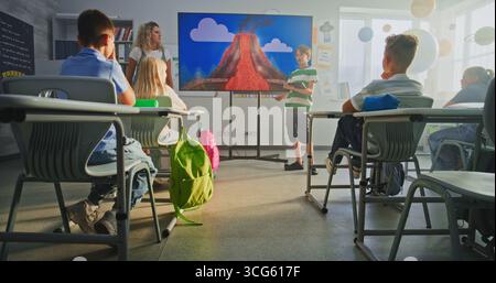 Grundschuljunge mit Tablet-Computer, der Kenntnisse der Geologie vor der Klasse und Lehrer mit digitalem Bildschirm vorstellt. Die Schüler hören sich die Präsentation an, der Lernprozess des Vulkanausbruchs. Stockfoto
