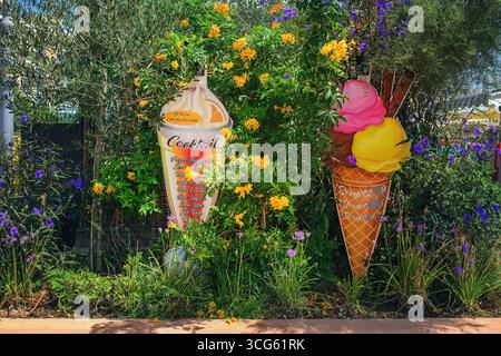 Helle Eis- und Cocktailschilder in Form von Waffelkegeln zwischen blühenden Pflanzen an einem sonnigen Tag. Stockfoto
