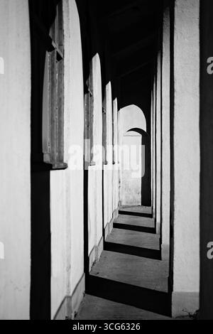 Schöne Schwarzweiß-Perspektive mit langen Bögen im Korridor. Sonnenlicht und kontrastreiche Schatten erzeugen ein hypnotisches abstraktes Hintergrundmuster in vertikaler Form Stockfoto
