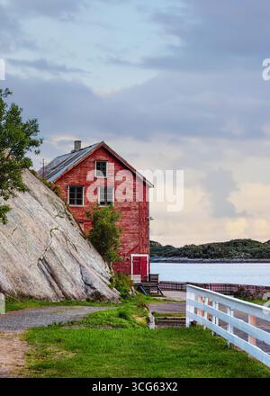 Rotes Holzhaus auf einer felsigen Klippe in der Nähe des ruhigen Wassers, umgeben von üppigem Grün und einem weißen Zaun, das ein malerisches Leben an der Küste zeigt. Lofoten Stockfoto