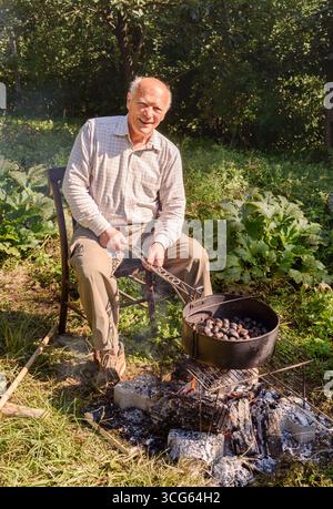 Älterer Mann, der frische Kastanien in einer Pfanne über heißen Kohlen auf einem Grill röstet. Stockfoto