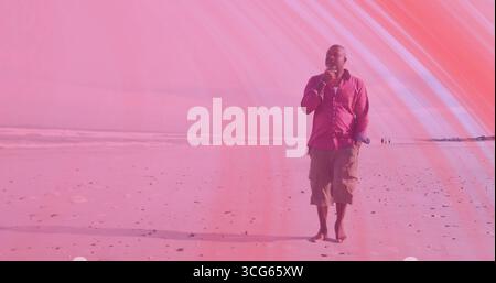 Ein älterer Mann in rosa Hemd und Shorts, der barfuß über den Strand fährt, mit Smartphone, Kopierraum Stockfoto