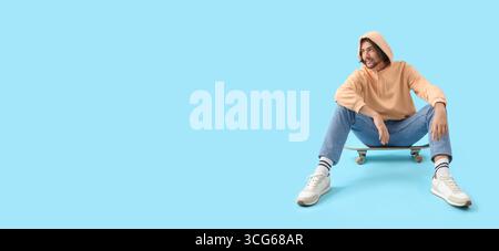 Junger Mann, der auf Skateboard vor blauem Hintergrund sitzt und Platz für Text hat Stockfoto