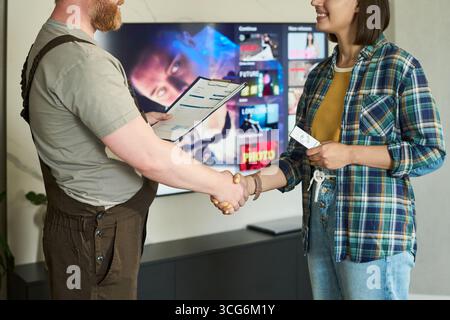 Kaukasischer Mann mittleren Alters im Arbeitsanzug schüttelt die Hand mit einer jungen, kaukasischen Frau, die eine Fernbedienung hält, beide stehen vor dem Fernseher und zeigen ein Streaming-Menü an Stockfoto