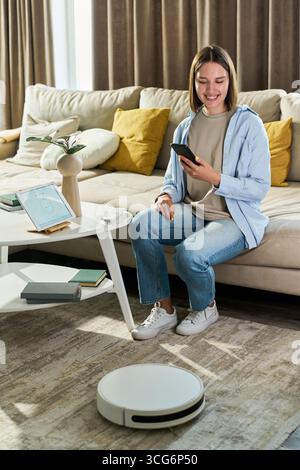Kaukasische junge Erwachsene Frau, die auf dem Sofa sitzt und mit dem Smartphone den Roboter-Staubsauger im Wohnzimmer steuert, lächelt und mit Smart Home interagiert Stockfoto