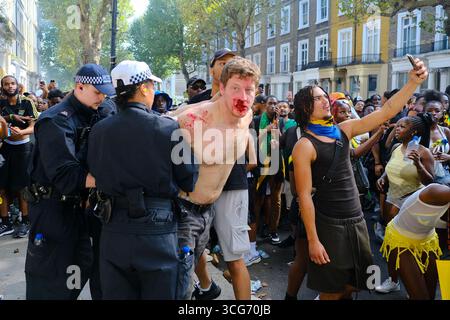 Ein Mann mit einem blutigen Mund wird nach einer Auseinandersetzung vor einem Haus während des Notting Hill Carnival verhaftet. Eine Menge versammelt sich, als ein Zuschauer ein Selfie mit ihm macht. Stockfoto