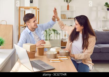 Junge glückliche Paare sitzen mit Essen zum Mitnehmen und geben sich gegenseitig High-Five. Stockfoto