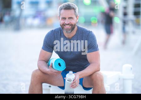 Sport-Fitness-Mann, der draußen auf einer Bank ruht. Reifes Fitnesssportmodell, das nach dem Training auf einer Bank sitzt. Senior Sports man macht danach eine Pause Stockfoto