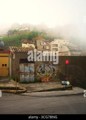 Nebelige Morgenstraße in Enna mit Graffiti an der alten Mauer, Bushalteschild und Hügelhäusern, die in Nebel verschwinden und eine stimmungsvolle sizilianische Stadtszene schaffen Stockfoto